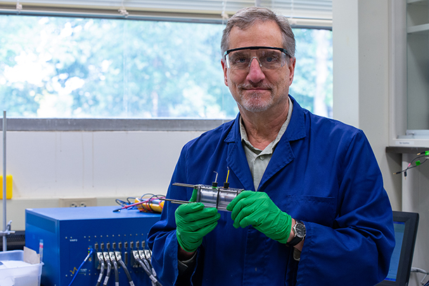 A man in a blue lab coat and protective glasses holds a small electrochemical reactor.