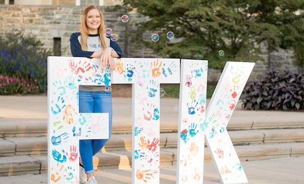 a woman stands by large white F T K letters covered with children's handprints in bright colors