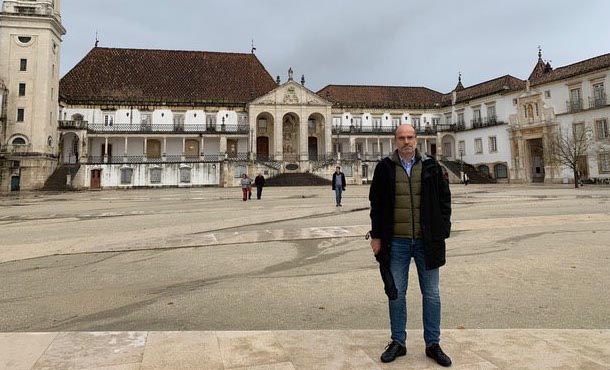 A man in jeans and a jacket stands before a rustic university building in Portugal.