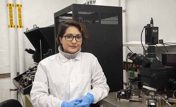 A woman in glasses and a lab coat poses in front of lab equipment.
