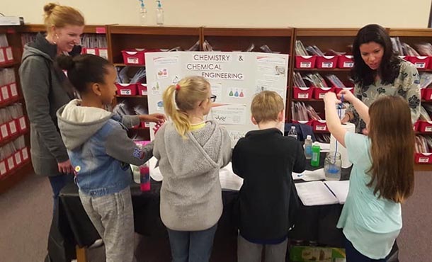 two women, three girls, and one boy review a poster at a science fair
