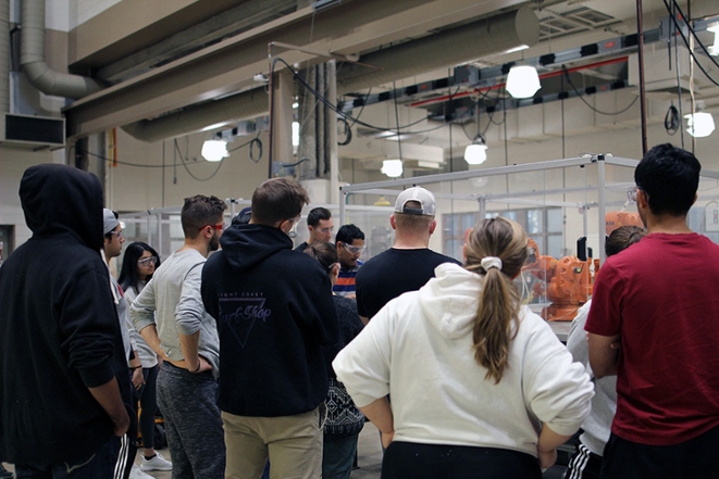 A large group of students stand around a robot in a glass encasement.