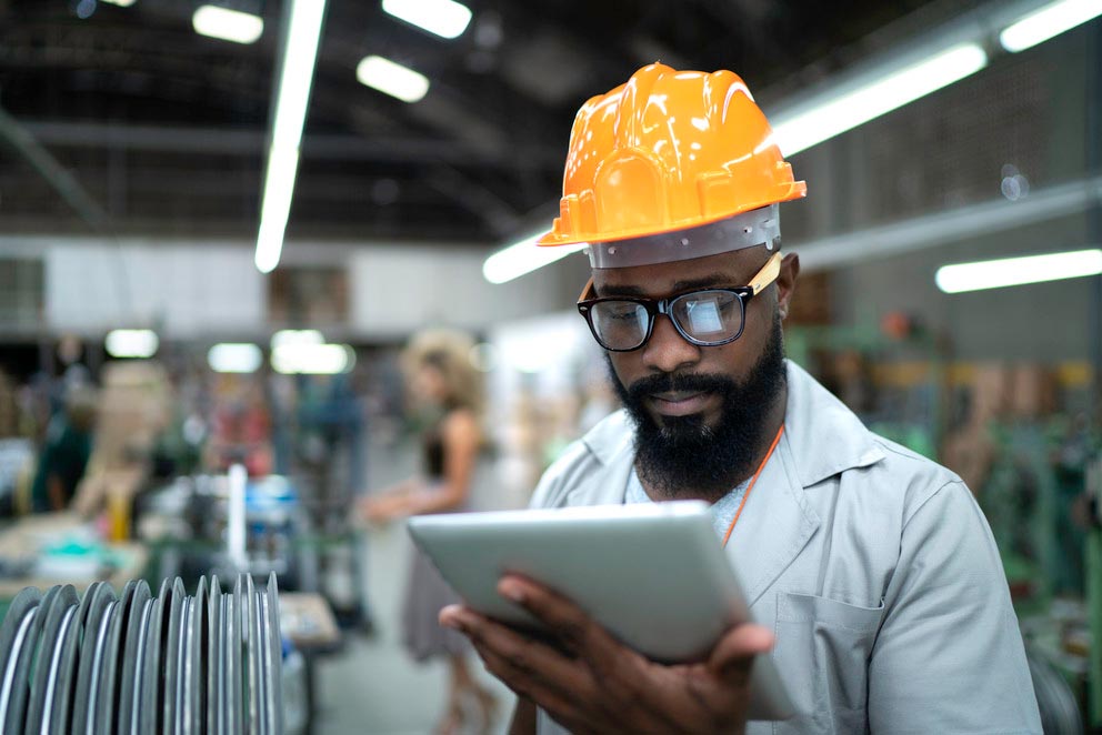 a man wearing a yellow hardhat looks at a tablet computer while standing in a manufacturing facility