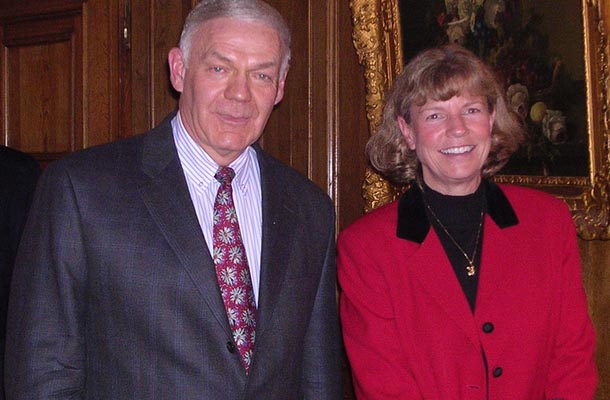 A man wearing a suit and tie and a woman in a red blazer stand and smile at the camera.