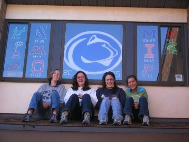 Four women sit on a rooftop with the Penn State Nittany Lion athletics logo painted on the window behind them
