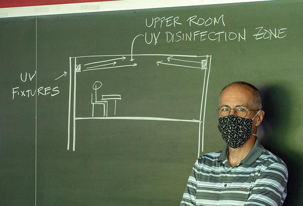 a man wearing a facemask and glasses points to a chalkboard with a classroom diagram with the words upper room UV disinfection zone and UV fixtures