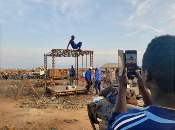 Image of a person taking a photo on their cell phone of four men around a metal structure in the middle of a scrapyard.