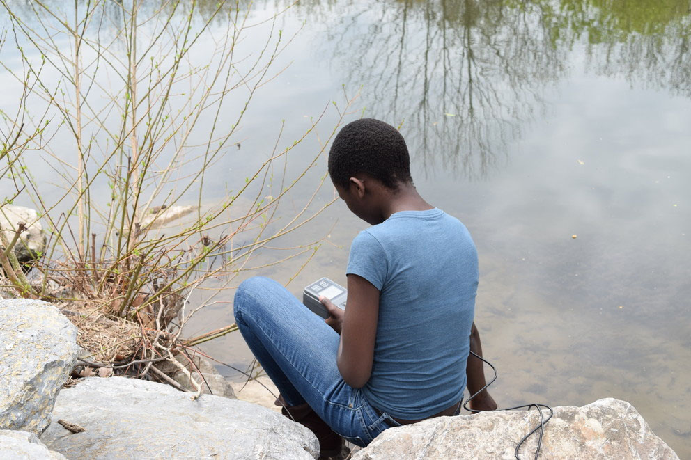 Woman sits by river basin with handheld meter.