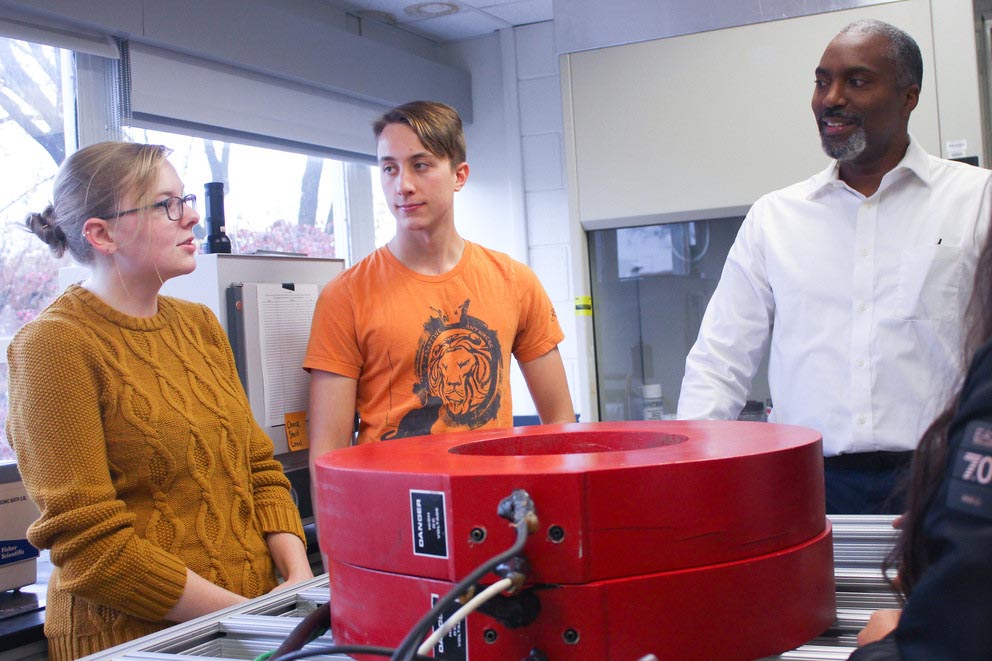 Two graduate students and a professor stand near a magnetic separation device, discussing their research.