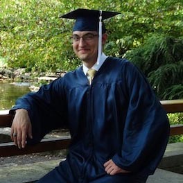 a man wearing a cap and gown poses on a foot bridge in a garden