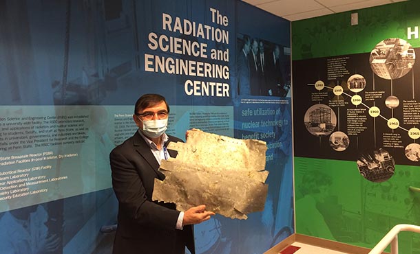 A man in a face mask and glasses stands in front of a blue wall that reads Radiation Science and Engineering Center holding a piece of metal with rough edges and rivet holes.