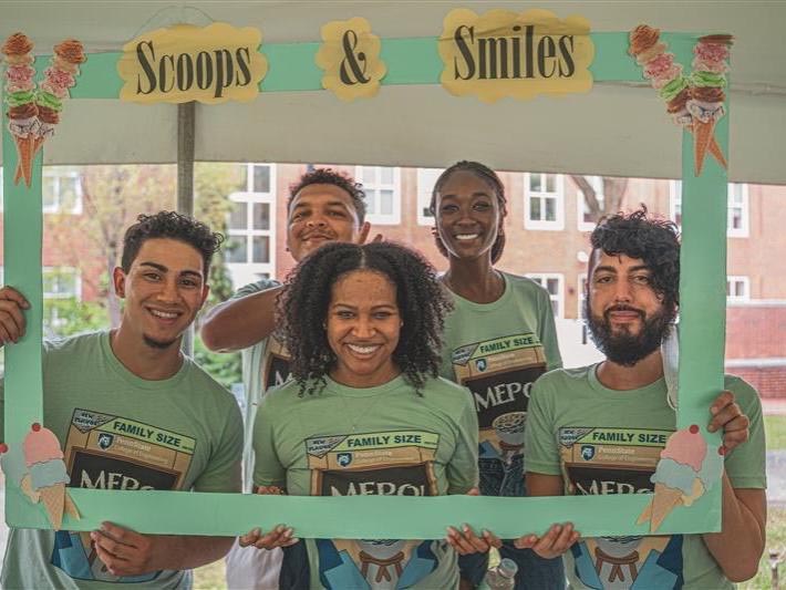 Five students pose behind a large cardboard photo frame that has the phrase scoops and smiles printed across the top
