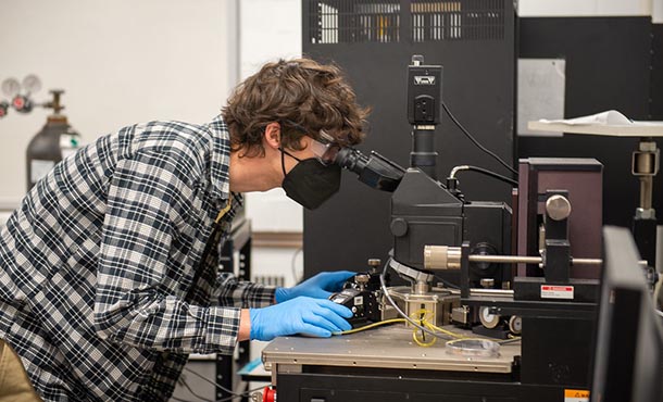 A person wearing a black face mask, blue gloves and a plaid shirt looks into a microscope. 