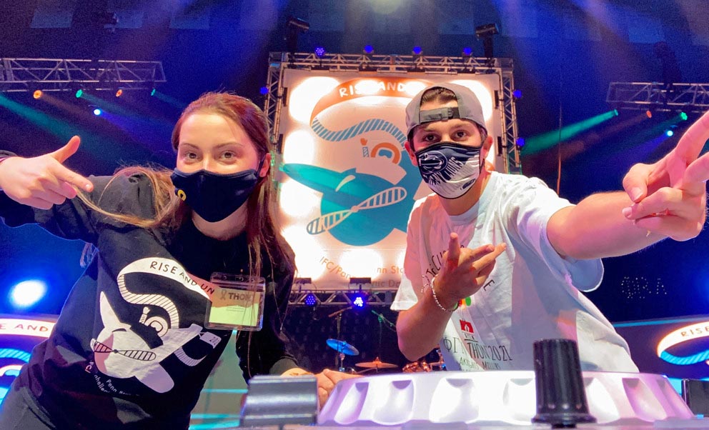 Two college students wearing face masks pose for the camera on a brightly lit stage.