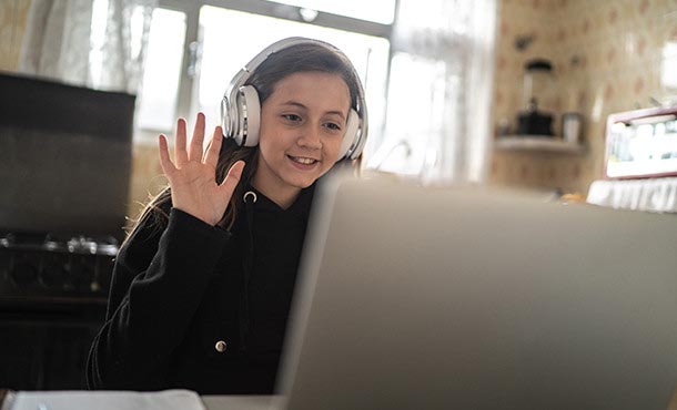 A girl wearing a sweatshirt and large headphones smiles and raises her hand in front of an open laptop computer.