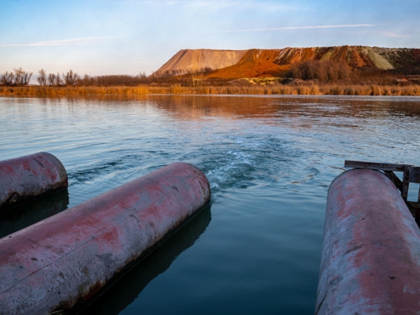 Three rusted pipes outputting into a lake with red and white hills in the distance. 