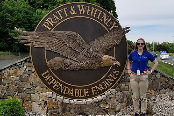 A female undergraduate student poses next to a sign that features the Pratt and Whitney logo.