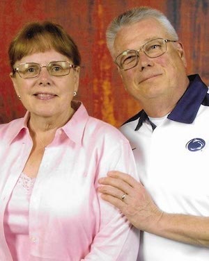 A woman in a pink shirt and glasses and a man in a white Penn State polo shirt with blue accent colors and glasses smile at the camera.
