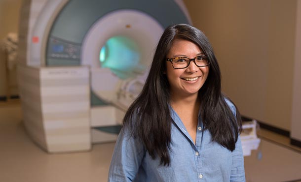 A young woman in a blue shirt and glasses stands in front of an MRI machine.