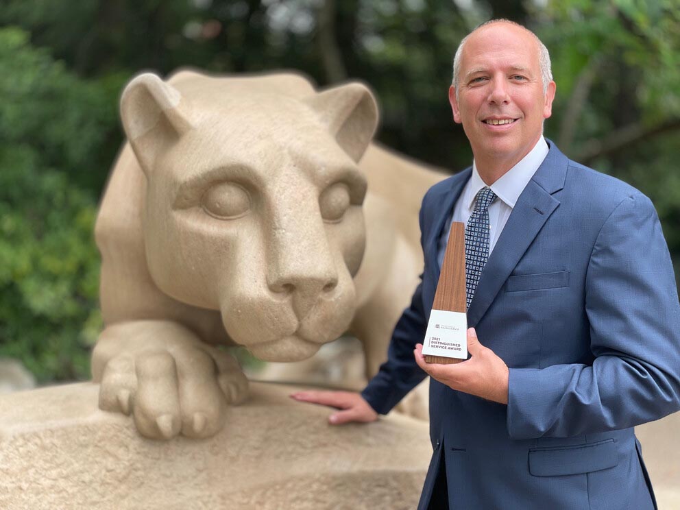 A man in a suit and tie smiles at the camera and holds a brown triangular award. The Nittany Lion statue is visible behind him.