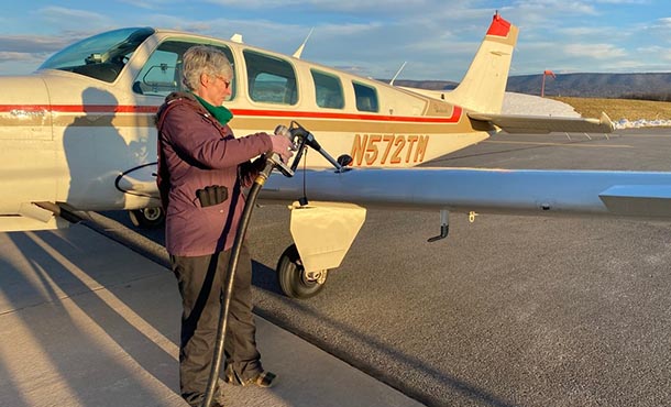 A woman holds a fuel pump to a small aircraft. A mountain is in the background.