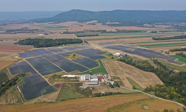An aerial view of solar panels surrounded by farmland and bar with mountains in the background.