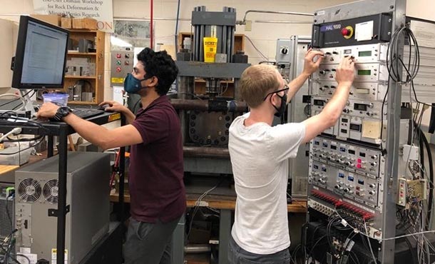 two students in an engineering lab, one student is on a computer and the other student is turning dials on a large machine