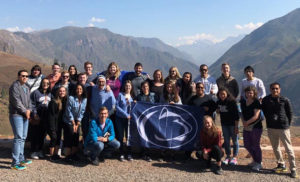 A group of 27 students and one professor pose in front of a mountain range while holding a Penn State flag