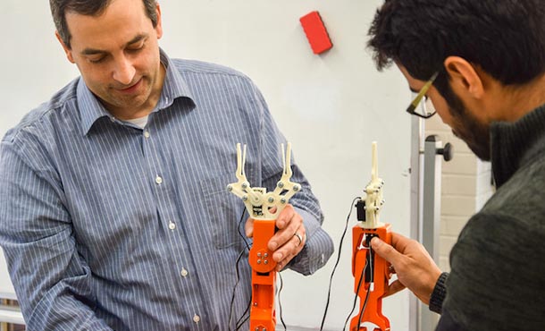 a professor on the left holds a robot arm, while a student on the right holds another robot arm