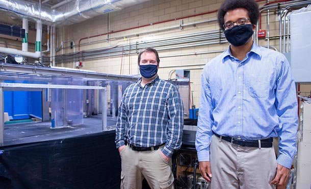 Two professors wearing face masks stand besides an experimental wind tunnel in a laboratory.