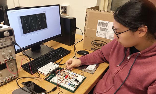 A student in a zip-up sweatshirt and glasses works on a circuit board in front of a computer screen displaying a graph