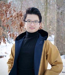 A person wearing glasses, a black turtle neck, and a yellow jacket poses outside on a winter day in front of some trees