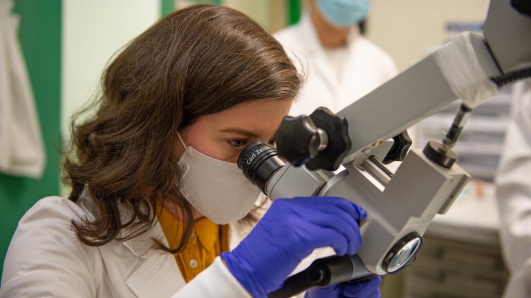 a close up photo a woman with brown hair, a lab coat and blue gloves looking into a microscope