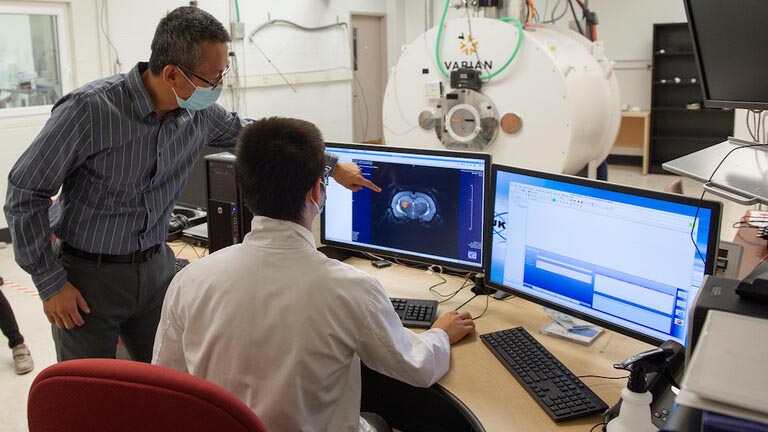 One man, standing, points to an image on a computer screen to a man, who is sitting at a desk. A large white tube is shown behind them