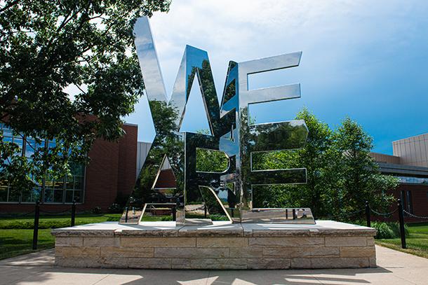 mirrored "we are" word sculpture against backdrop of brick building and blue sky