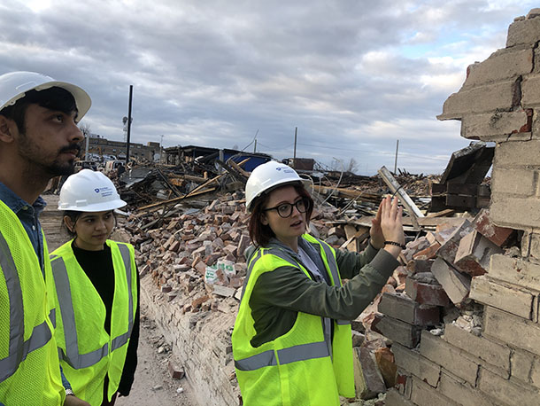 Three people wearing hard hats and neon vests stand near a mostly demolished brick wall as one of the people gestures at the wall in explanation to the other two people who look on. 