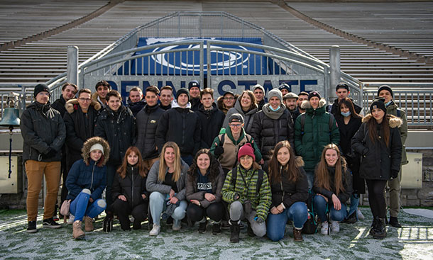 A group of people wearing winter coats poses in front of metal gates that say 'Penn State.'