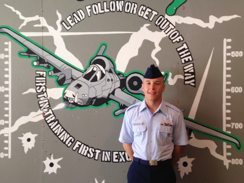 A person wearing Air Force blues stands with hands behind his back in front of a large illustration of an airplane.