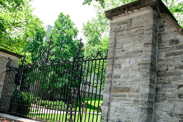 An iron gate stretches between two stone pillars. Green grass and trees are visible beyond the gate.