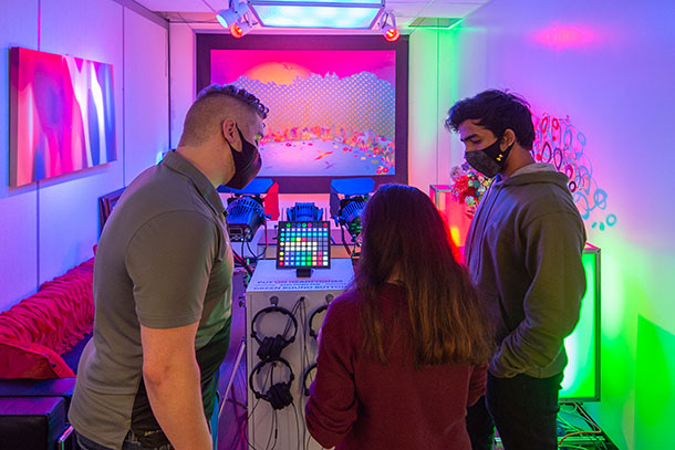 Three people study a display screen in a technical lab with brightly colored lights.