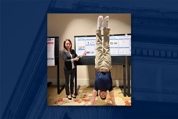 A person does a handstand in front of a poster while another person gives a thumbs up