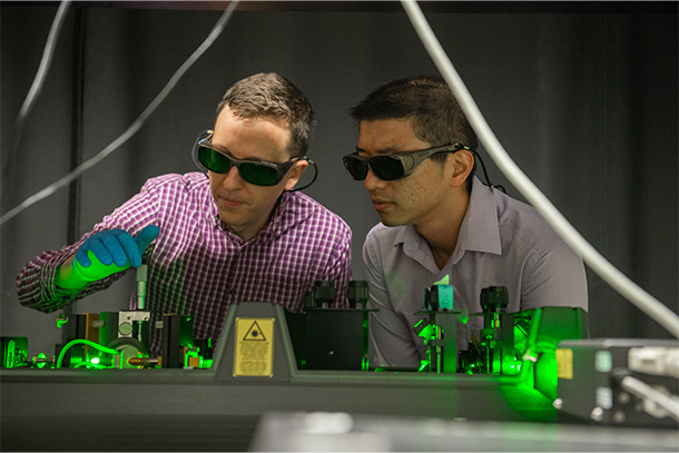 Two people wearing safety glasses lean over a table holding a laser shining a green light.