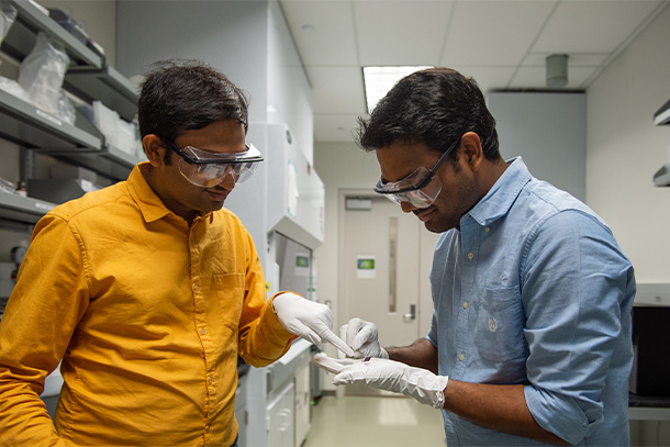 Two individuals wearing lab glasses and gloves in a lab point to a small computer chip