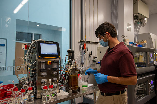 A man in a maroon polo shirt and a blue face mask works at a lab table with containers of liquids, many wires and a small display screen.