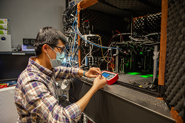 A researcher in a plaid shirt and blue face mask adjusts a set of wires in a lab setting.