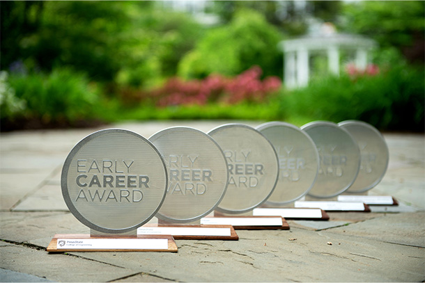  Six awards are lined up on stone, with greenery in the background. Each Award is a silver circle erected on a wood stand.