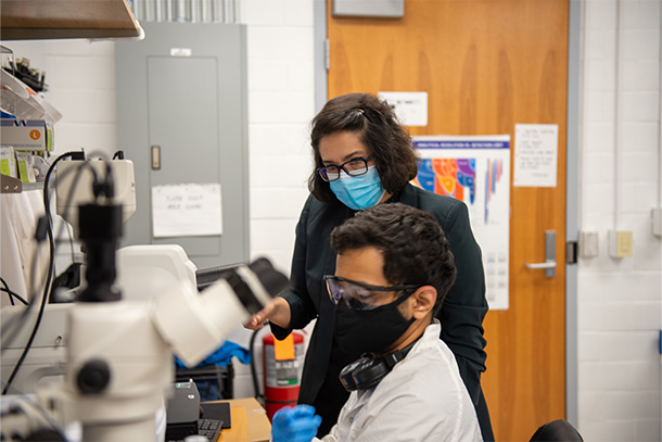 Dark-haired person wearing black suit, glasses and mask stands next to dark-haired person with mask and white lab coat seated at microscope in lab.