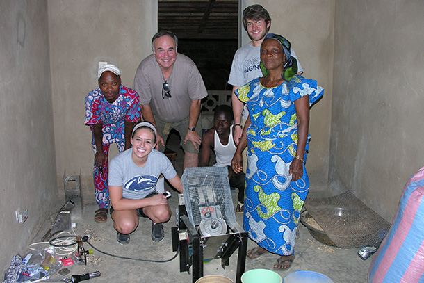 A group of people stand around a small silver and black machine with a mesh overlay in a brown room. Bowls full of baobab seeds are on the floor. 