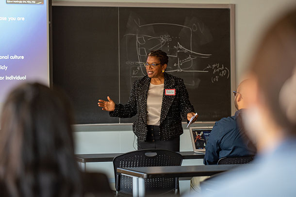 A woman in a blazer speaks in front of a chalkboard and projection screen while students in the foreground look on. 