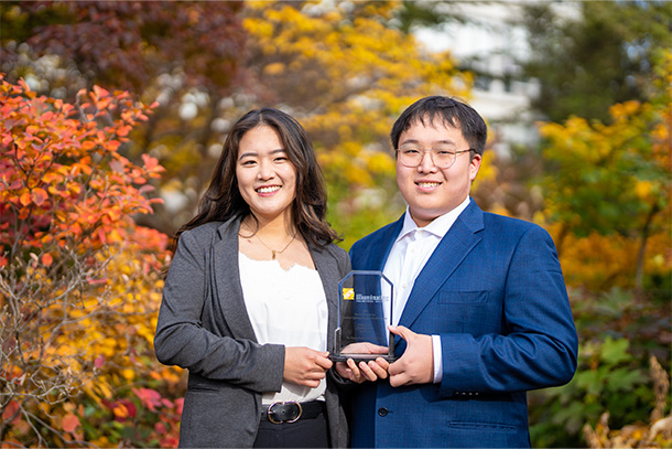 two people in business suits hold crystal trophy in front of backdrop of fall leaves